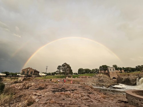 A rainbow arches over a rocky landscape with a waterfall, while people walk near historic buildings.