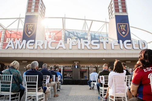 A crowd gathers at America First Field for an event, with a speaker visible in front of the stadium's entrance.