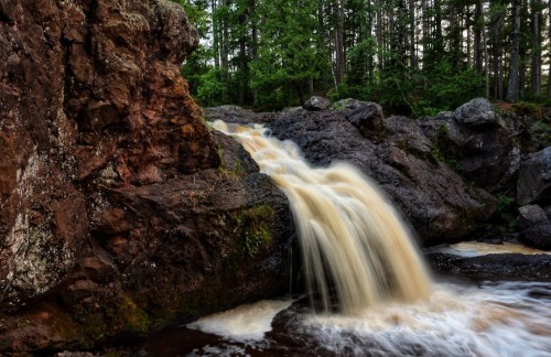 A small waterfall cascades over rocky terrain, surrounded by lush green trees in a serene forest setting.
