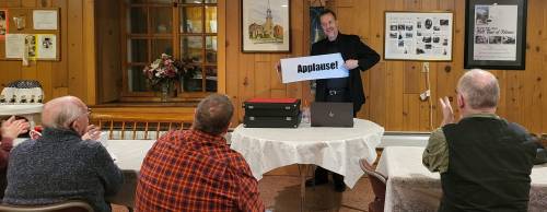 A speaker holds an "Applause!" sign while an audience claps at a gathering in a wooden-paneled room.