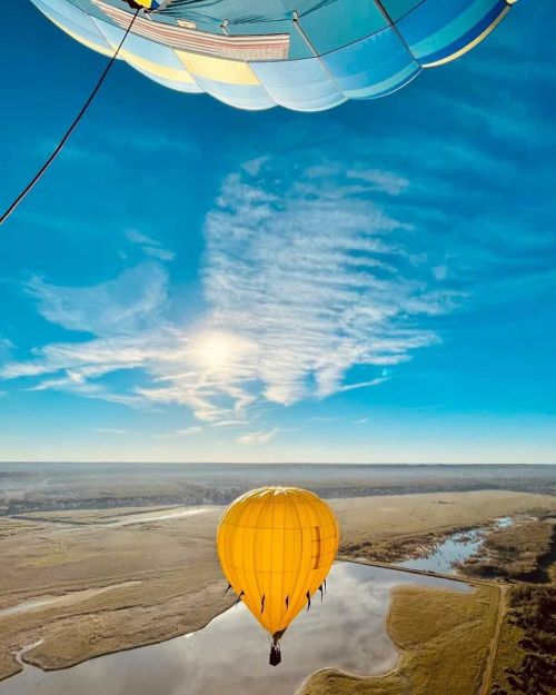 A vibrant yellow hot air balloon floats below a colorful balloon against a clear blue sky and scenic landscape.