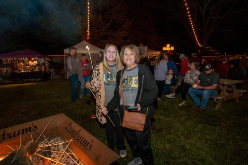 Two women smile by a bonfire, roasting marshmallows, with a festive crowd and lights in the background.