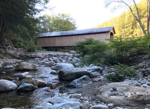 A wooden covered bridge beside a rocky stream, surrounded by trees and greenery in a serene natural setting.