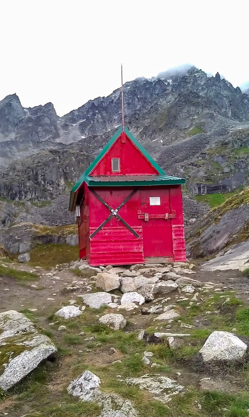 A small red cabin with a green roof sits among rocky terrain and mountains under a cloudy sky.