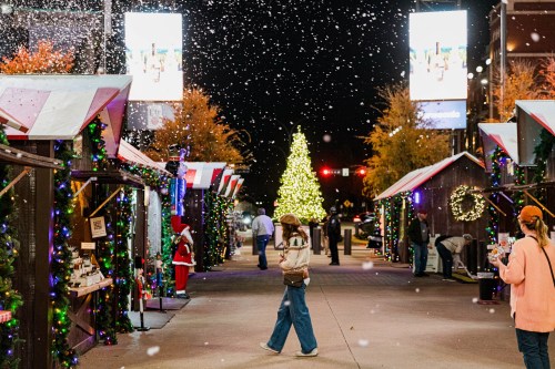 A festive street scene with holiday decorations, snow falling, and a large Christmas tree in the background.