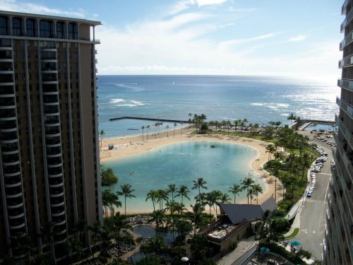 A scenic view of a beach lagoon surrounded by palm trees, with ocean waves and buildings in the background.