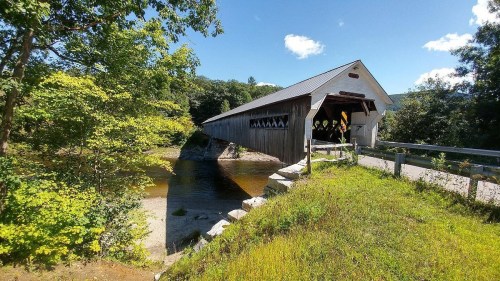 A scenic view of a covered bridge over a calm river, surrounded by lush greenery and a clear blue sky.