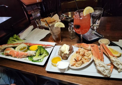 A table set with two plates of seafood, including crab legs, shrimp, broccoli, and drinks, in a cozy restaurant setting.