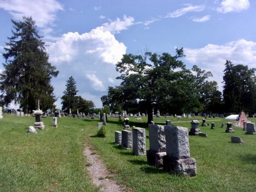 A peaceful cemetery with gravestones scattered across a grassy area, under a blue sky with fluffy clouds.