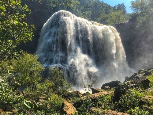 A majestic waterfall cascades over rocky cliffs, surrounded by lush greenery and a clear blue sky.