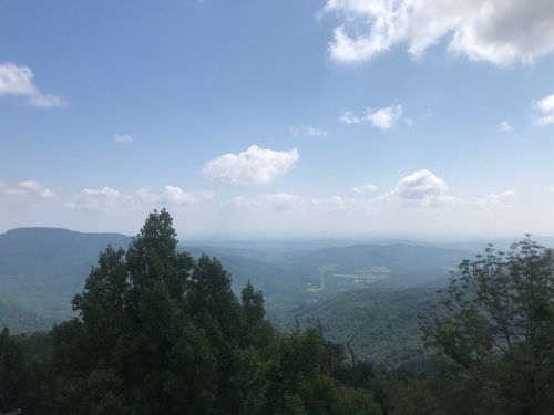 A scenic view of rolling mountains under a blue sky with fluffy clouds, framed by green trees in the foreground.