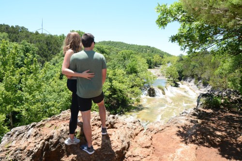 A couple stands on a rocky ledge, overlooking a lush green landscape and a flowing river with a wind turbine in the distance.