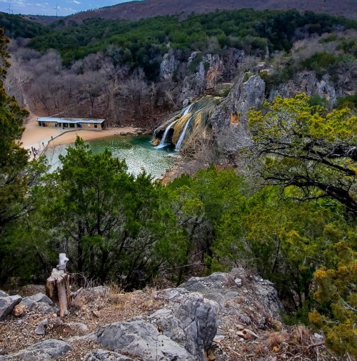 A scenic view of a waterfall cascading into a green pool, surrounded by rocky cliffs and lush trees.