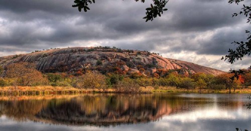 A rocky hill reflects in a calm lake, surrounded by trees and under a cloudy sky.