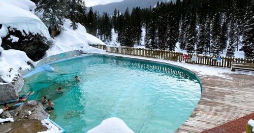 A scenic outdoor hot spring pool surrounded by snow-covered trees and mountains, with people enjoying the water.