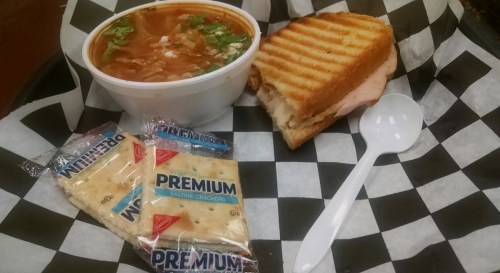 A bowl of soup, a grilled sandwich, and two packets of saltine crackers on a checkered tray.
