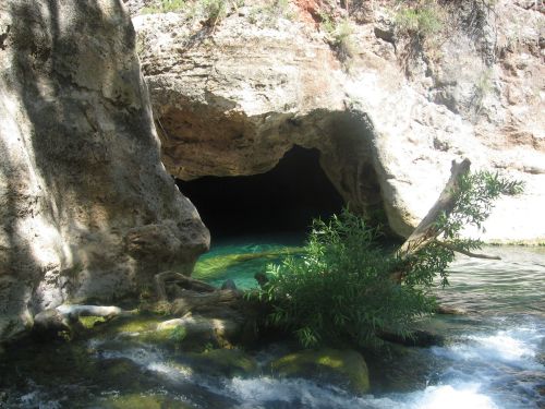 A serene cave entrance by a clear blue stream, surrounded by rocky terrain and lush greenery.