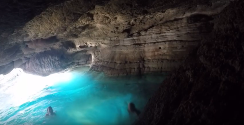 A serene cave with turquoise water, featuring two people swimming in the foreground. Natural rock formations surround the scene.