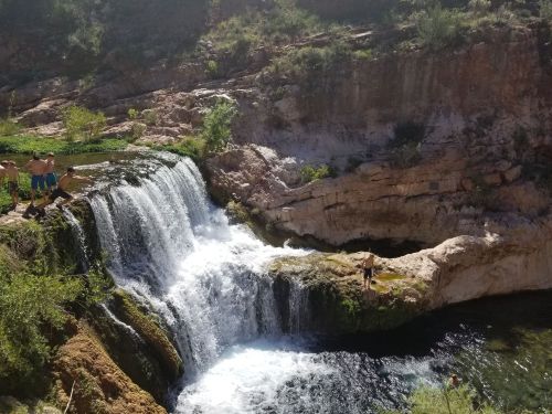 A scenic waterfall cascades into a pool, surrounded by rocky terrain and lush greenery. People enjoy the area nearby.