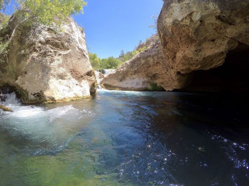 A serene river flows between rocky cliffs under a clear blue sky, with a small waterfall in the background.