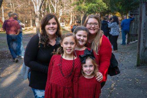 A group of five women and girls in matching red plaid outfits, smiling together outdoors in a park setting.