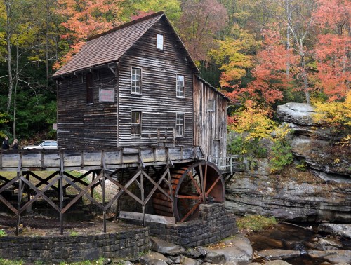 A rustic wooden mill with a water wheel, surrounded by colorful autumn foliage and rocky terrain.