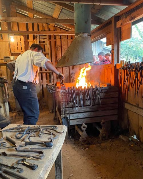 A blacksmith works at a forge, shaping metal as flames rise, surrounded by tools in a rustic workshop.