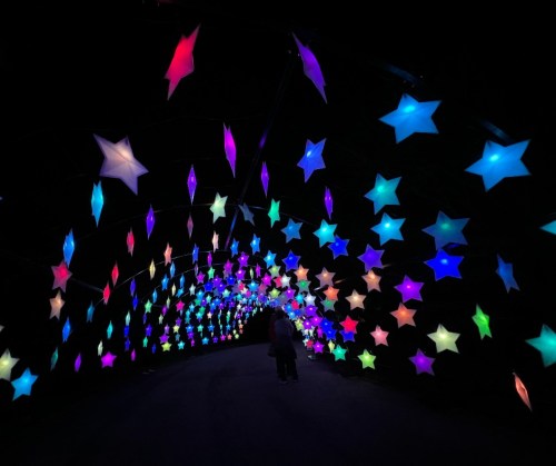 A colorful tunnel of illuminated stars in various shapes and colors against a dark background, with a person walking through.