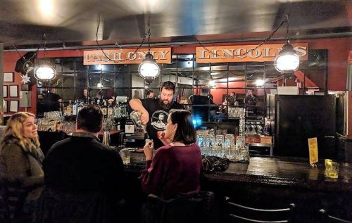 A bartender serves drinks to patrons at a cozy bar with warm lighting and a mirrored backdrop.