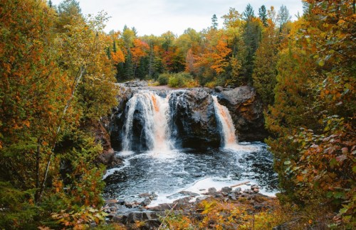 A scenic waterfall surrounded by vibrant autumn foliage and a calm river below.