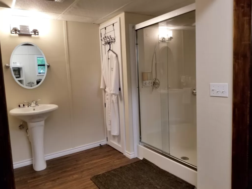 A modern bathroom featuring a shower, sink, mirror, and a robe hanging on the wall. Wood-like flooring is visible.