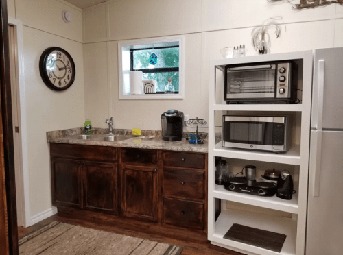 A cozy kitchen featuring a clock, countertop appliances, and a white shelving unit with a microwave and coffee maker.