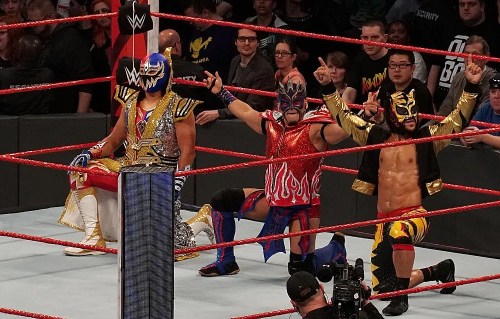 Three masked wrestlers pose in a wrestling ring, surrounded by an enthusiastic crowd. Red ropes frame the scene.