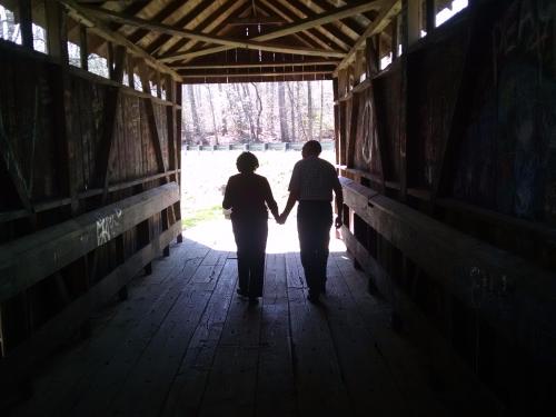 A couple holding hands walks through a covered bridge, silhouetted against bright light at the exit.