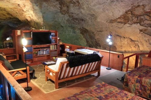 Cozy cave interior with wooden furniture, a TV, bookshelves, and colorful bedding, illuminated by lamps.