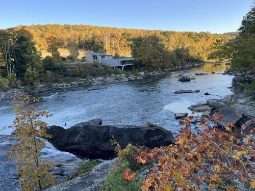 A serene river flows past rocky banks, surrounded by autumn foliage and a modern building in the background.