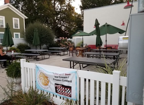 Outdoor seating area at an ice cream shop, featuring tables, umbrellas, and a banner promoting their frozen custard.