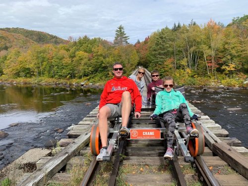 A family enjoys a scenic ride on a rail bike along a river, surrounded by colorful autumn foliage.