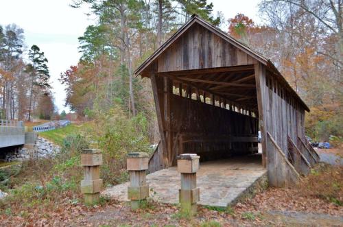 A rustic wooden covered bridge stands beside a road, surrounded by autumn foliage and trees.