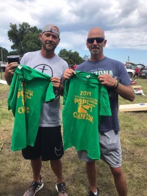 Two men hold up green t-shirts that read "2017 Popcorn Festival Champions" while standing outdoors.