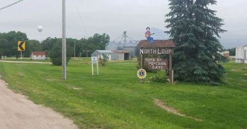 Sign for North Loup, Nebraska, noting it as the "Home of Popcorn Days," with rural scenery and buildings in the background.