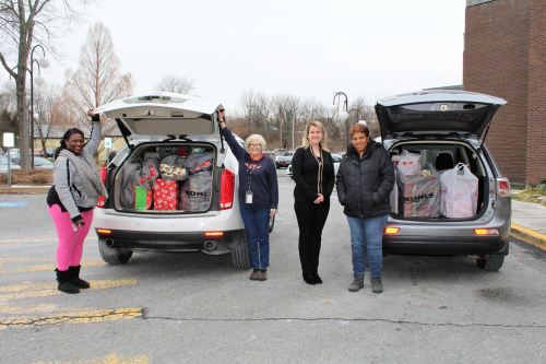 Four women stand by two cars with open trunks filled with wrapped gifts, smiling in a parking lot.