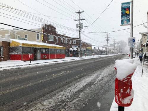 A snowy street scene with a red and yellow restaurant, power lines, and a snow-covered sidewalk.