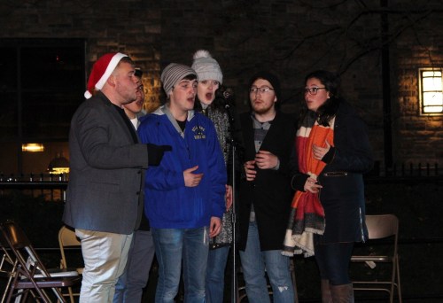 A group of five singers performs outdoors at night, wearing winter clothing and festive accessories.