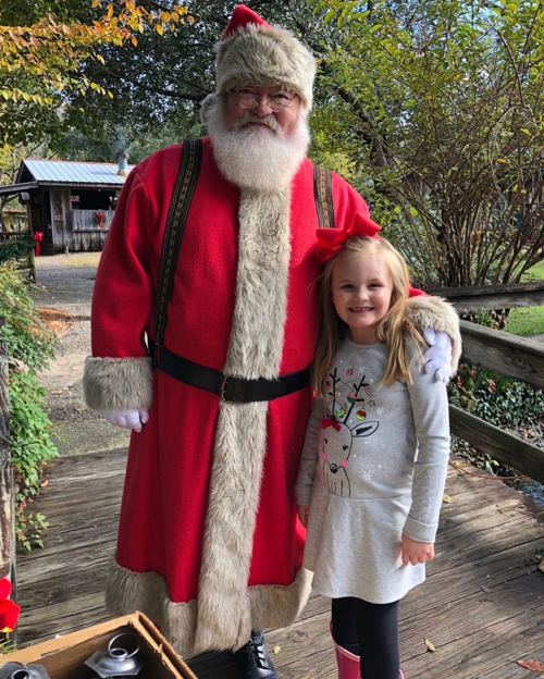 A smiling girl in a reindeer shirt stands next to Santa Claus in a festive setting.