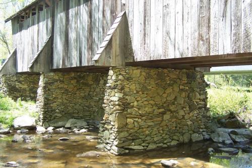 A wooden covered bridge supported by stone pillars over a shallow stream, surrounded by greenery.