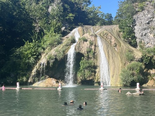 A serene waterfall cascades into a green pool, with people swimming and enjoying the natural surroundings.