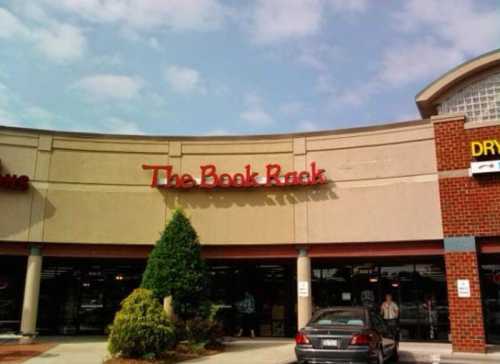 Exterior of a bookstore called "The Book Rack," featuring a sign and nearby greenery under a partly cloudy sky.