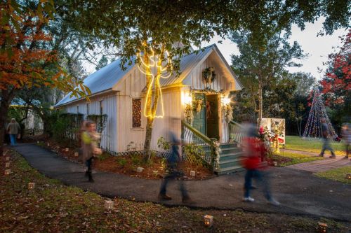 A charming white house decorated with holiday lights, surrounded by trees and people walking along a path.