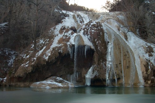 A frozen waterfall cascading over rocky terrain, surrounded by snow and trees, reflecting in a calm pool below.